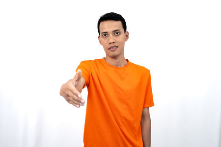 Portrait of a young Asian man wearing an orange t-shirt posing giving a warm greeting to a friend or customer. Isolated on white background.の写真素材