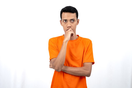 Portrait of a young Asian man wearing an orange t-shirt posing thinking, searching for ideas, or confused. Isolated on white background.の写真素材