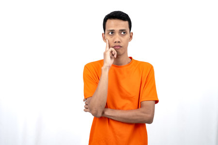 Portrait of a young Asian man wearing an orange t-shirt posing thinking, searching for ideas, or confused. Isolated on white background.の写真素材