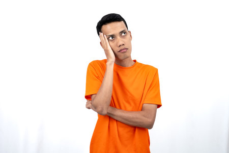 Portrait of a young Asian man wearing an orange t-shirt posing thinking, searching for ideas, or confused. Isolated on white background.の写真素材