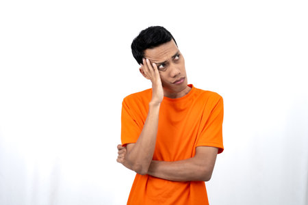 Portrait of a young Asian man wearing an orange t-shirt posing thinking, searching for ideas, or confused. Isolated on white background.の写真素材