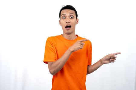 Portrait of a young Asian man wearing an orange t-shirt pointing aside copy space for promotional space. isolated on white background.の写真素材