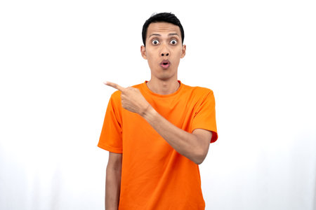 Portrait of a young Asian man wearing an orange t-shirt pointing aside copy space for promotional space. isolated on white background.の写真素材