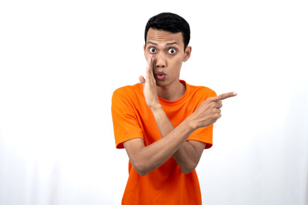 Portrait of a young Asian man wearing an orange t-shirt with his hand near his mouth in a whispering pose while pointing to the side of the copy space. Isolated on white background.の写真素材