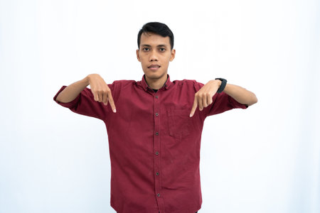 Asian man, businessman or student concept wearing red casual shirt pointing down copy space with happy or surprised expression. Isolated white background.の写真素材