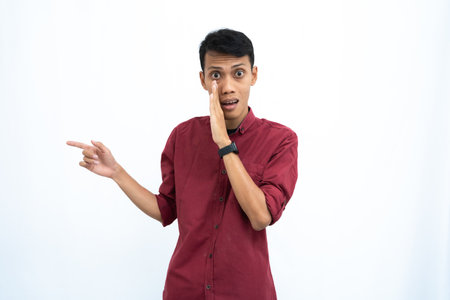 Asian man, wearing red casual shirt, pointing sideways, whispering with hand in mouth, giving information, isolated on white background.の写真素材