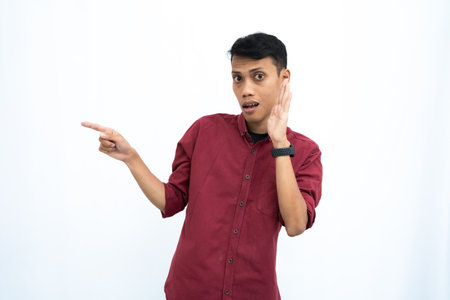 Asian man, wearing red casual shirt, pointing sideways, whispering with hand in mouth, giving information, isolated on white background.の写真素材