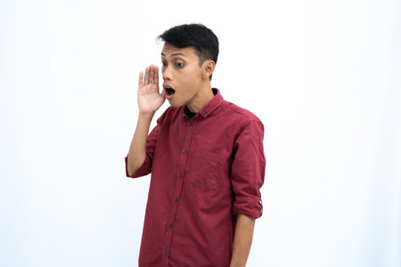 Asian man, businessman or student concept wearing red casual shirt, shouting with hand in mouth, giving information or whispering, isolated on white background.の写真素材