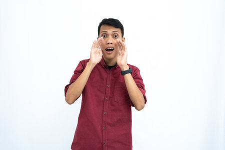 Asian man, businessman or student concept wearing red casual shirt, shouting with hand in mouth, giving information or whispering, isolated on white background.の写真素材