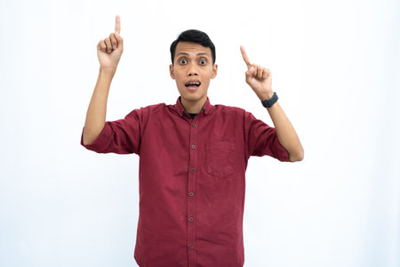 Asian man, businessman or student concept wearing red casual shirt pointing up at the copy space with a happy or surprised look on his face. Isolated white background.の写真素材