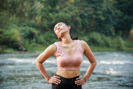 A slender, tanned girl in sportswear is doing neck roll stretching exercises in the open air on the edge of a beautiful river, before exercising.の写真素材