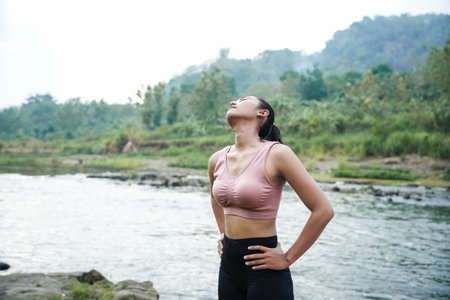A slender, tanned girl in sportswear is doing neck roll stretching exercises in the open air on the edge of a beautiful river, before exercising.の写真素材