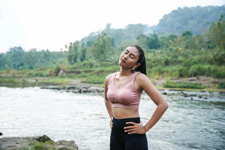 A slender, tanned girl in sportswear is doing neck roll stretching exercises in the open air on the edge of a beautiful river, before exercising.の写真素材