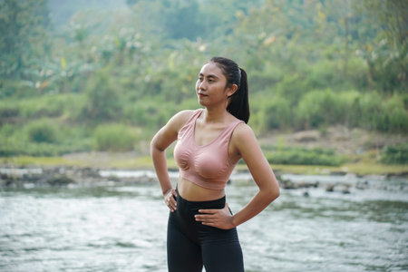 A slender Asian girl with brown skin in sportswear is doing hip circle stretching movements in the open air on the edge of a beautiful river, before doing sports.の写真素材