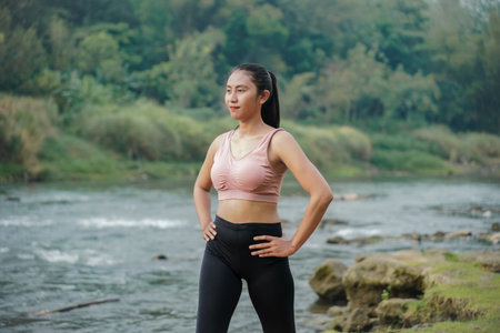 A slender Asian girl with brown skin in sportswear doing stretching movements and rotating her body in the open air on the edge of a beautiful river, before doing sports.の写真素材