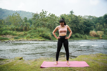 A slender, tanned girl in sportswear is doing neck roll stretching exercises in the open air on the edge of a beautiful river, before exercising.の写真素材