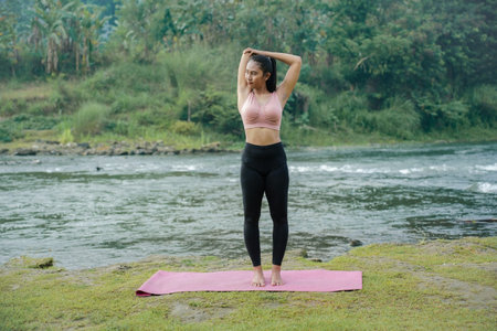 A slender Asian girl with brown skin in sportswear is doing tricep stretching movements in the open...
