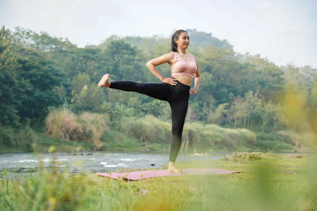 A slender Asian girl with brown skin in sportswear doing stretching movements and swinging her legs in the open air on the edge of a beautiful river, before doing sports.の写真素材