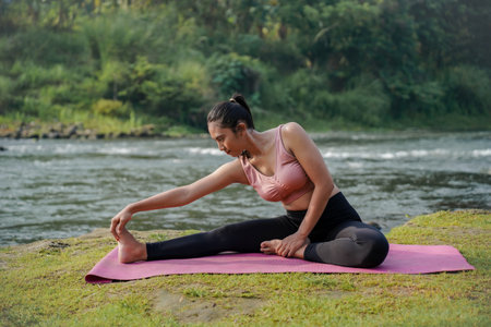 A slender Asian girl with brown skin in sportswear is doing stretching movements of her hamstrings in the open air on the edge of a beautiful river, before doing sports.の写真素材