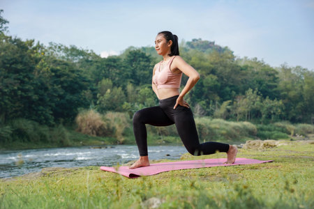 A slender, tanned Asian girl in sportswear performs lunge stretching exercises outdoors in a...