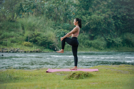 A slender Asian girl with brown skin in sportswear doing muscle warm-up movements in the open air on the edge of a beautiful river, before doing sports.の写真素材