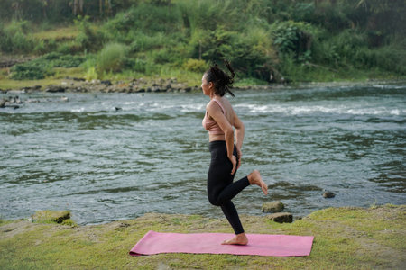 A slender, tanned Asian girl in sportswear performs a stretch in the open air by a beautiful...