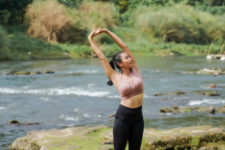 Mental and physical health. A slender, tanned Asian girl in sportswear does yoga by a calm river in the morning. The woman stands in Tadasana pose with her hands above her head.の写真素材