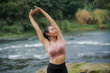 Mental and physical health. A slender, tanned Asian girl in sportswear does yoga by a calm river in the morning. The woman stands in Tadasana pose with her hands above her head.の写真素材