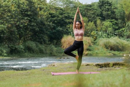 Mental and physical health. A slender, tanned Asian girl in sportswear does yoga by a calm river in the morning. The woman stands in Vrksasana pose with her hands above her head.の写真素材