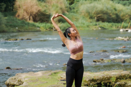 Mental and physical health. A slender, tanned Asian girl in sportswear does yoga by a calm river in...