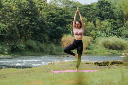 Mental and physical health. A slender, tanned Asian girl in sportswear does yoga by a calm river in the morning. The woman stands in Vrksasana pose with her hands above her head.の写真素材