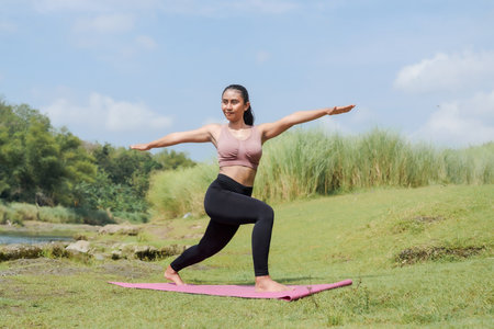 Mental and physical health. A slender, tanned Asian girl in sportswear does yoga by a calm river in the morning. The woman stands in a warrior pose with her arms outstretched.の写真素材