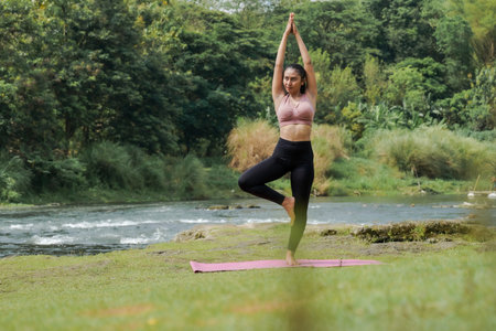 Mental and physical health. A slender, tanned Asian girl in sportswear does yoga by a calm river in the morning. The woman stands in Vrksasana pose with her hands above her head.の写真素材