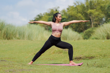 Mental and physical health. A woman in sportswear does yoga by a calm river in the morning. The woman stands in a warrior pose with her arms outstretched.の写真素材