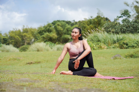 Mental and physical health. A slender, tanned Asian woman in sportswear performs yoga outdoors in a beautiful riverbank in the morning. The woman is performing Ardha Matsyendrasana, or Twist Pose.の写真素材