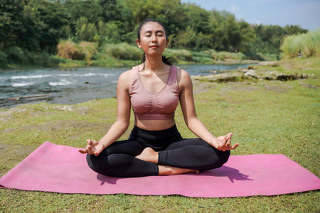 Mental and physical health. A slender, tanned Asian woman in sportswear practices yoga by a tranquil...