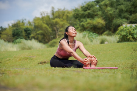 Mental and physical health. A slender, tanned woman in sportswear practices yoga by a tranquil river in the morning. The woman is performing Paschimottanasana, or Seated Forward Bend.の写真素材