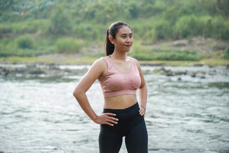 A slender woman with brown skin in sportswear doing stretching movements and rotating her body in the open air on the edge of a beautiful river, before doing sports.の写真素材