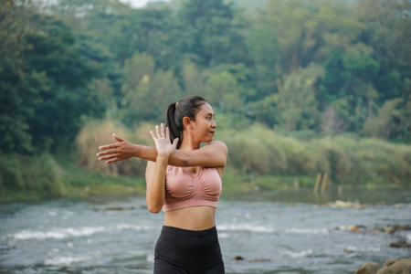 A slender Asian girl with brown skin in sportswear is doing shoulder muscle stretching movements in the open air on the edge of a beautiful river, before doing sports.の写真素材