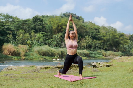Mental and physical health. A slender, tanned Asian girl in sportswear does yoga by a calm river in the morning. The woman stands in a warrior pose with her arms outstretched.の写真素材