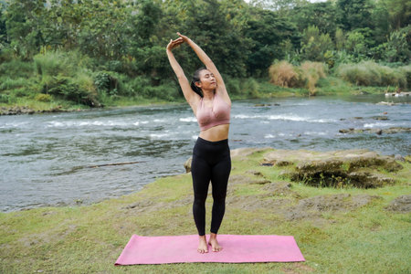 Mental and physical health. A slender, tanned Asian girl in sportswear does yoga by a calm river in...