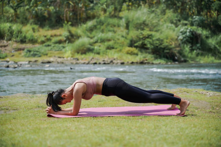 Mental and physical health. A slender, tanned Asian woman in sportswear practices yoga by a tranquil...