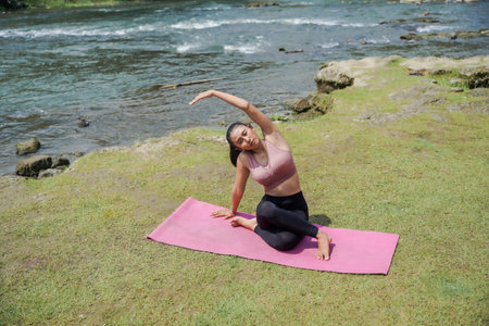 A slender, tanned Asian girl in sportswear performs yoga poses outdoors in a beautiful riverside...