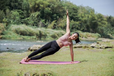 A slender, tanned Asian girl in sportswear performs yoga poses outdoors in a beautiful riverside...