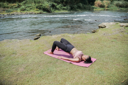 A slender, tanned girl in sportswear performs yoga poses outdoors in a beautiful riverside setting in the morning. The theme is meditation on health, mental, and physical well-being.の写真素材