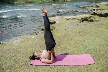 Mental and physical health. A slender, tanned Asian woman in sportswear performs yoga outdoors in a beautiful riverbank early in the morning. The woman is performing Sarvangasana, or Shoulder Stand.の写真素材