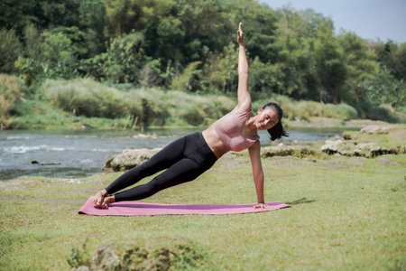 A slender, tanned Asian girl in sportswear performs yoga poses outdoors in a beautiful riverside...