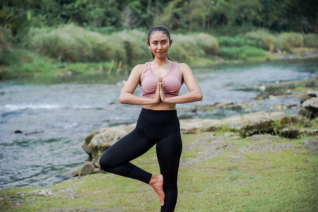 Mental and physical health. A slender, tanned Asian woman in sportswear performs yoga outdoors in a beautiful riverside setting in the morning. The woman is in Vrksasana, or Tree Pose.の写真素材