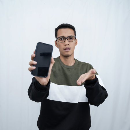 A young Asian man with glasses wearing a sweater points to a smartphone screen. Isolated on a white...