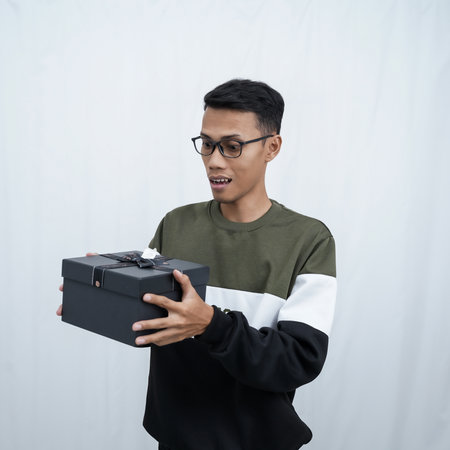 An Asian young man with glasses wearing a sweater with a surprised happy expression getting a surprise gift. Isolated on a white background.の写真素材